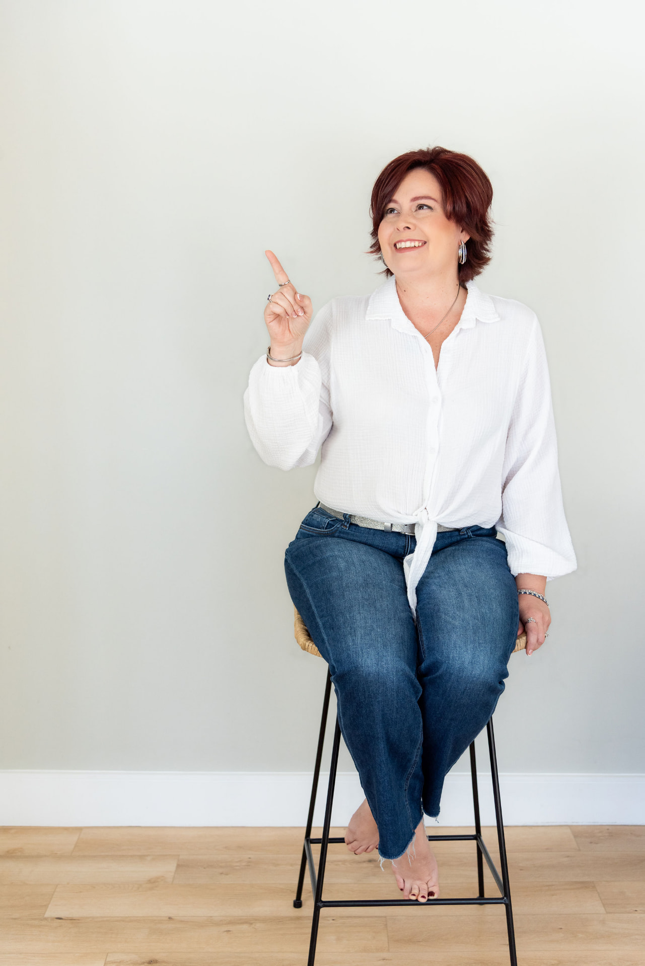 Melanie Rhora, keynote speaker and author, in a white blouse on a stool, pointing toward the From Good Girl to Goddess keynote description